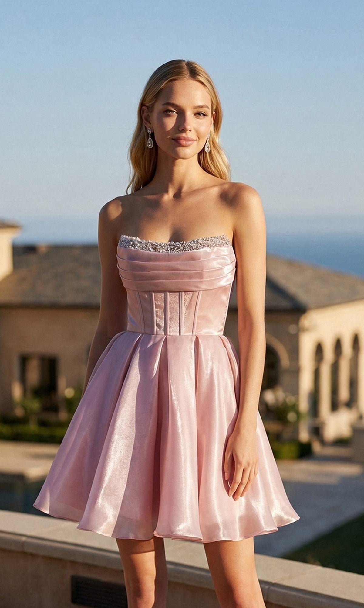 A woman stands outdoors in daylight, wearing the blush Juliet JT7187S short homecoming dress, featuring a strapless shimmer organza design with a fitted corset bodice and flared skirt. She pairs the look with light earrings and loose hair, with buildings visible in the background.